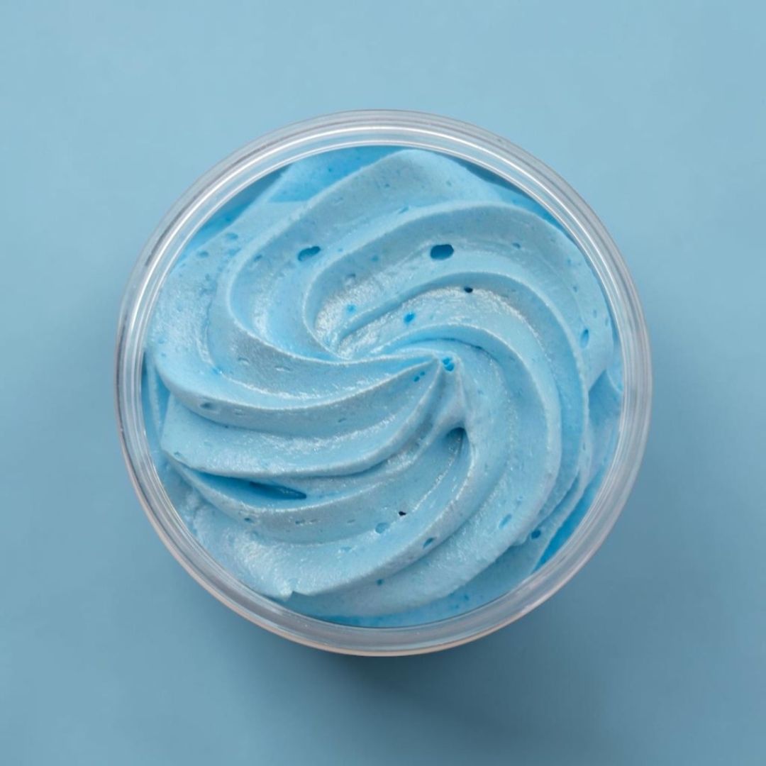A top-down view of light blue whipped soap swirled inside a clear plastic round container, set against a solid light blue background.