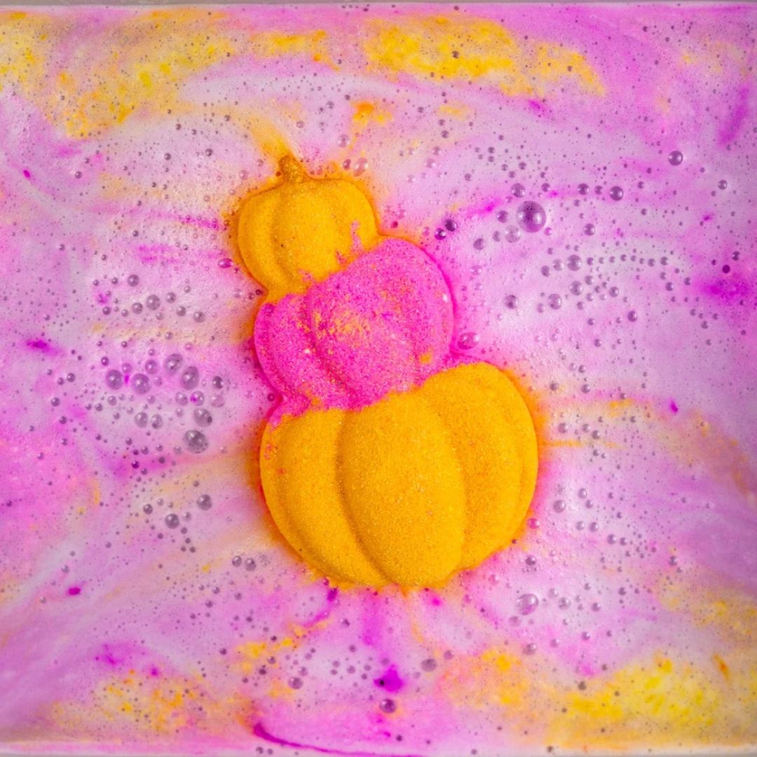 A close-up of a bath bomb shaped like a pumpkin, bursting in bright yellow and pink foam with bubbles on a pink and yellow marbled background.