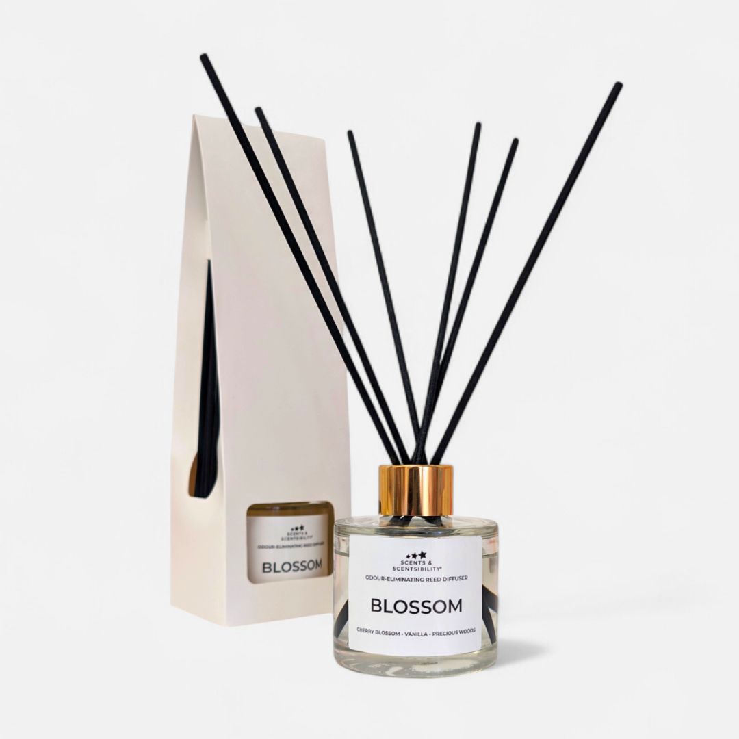 A glass bottle with clear liquid, gold collar and black fibre reeds with a white label with a presentation box in the background