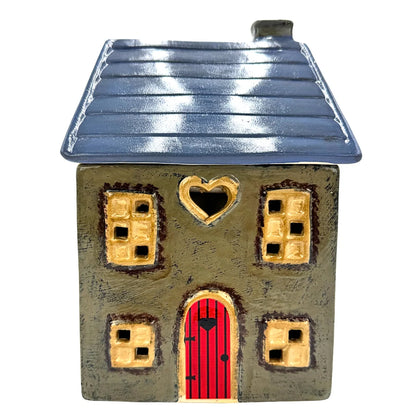 A ceramic cottage house wax melt warmer with a textured gray-brown body, a dark blue-gray roof, a bright red door, and gold-colored window and heart-shaped trim details, isolated on a white background.