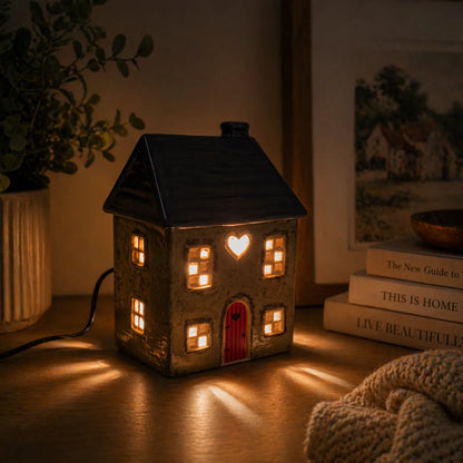 Ceramic house-shaped wax melt lamp with warm glow on a wooden surface, surrounded by books and a plant.
