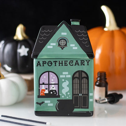 Green apothecary house-shaped wax melt & oil burner with pumpkins and bottles in the background