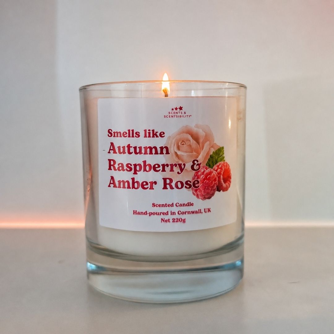 A lit candle in a glass jar labelled 'Autumn Raspberry & Amber Rose' on a light background.