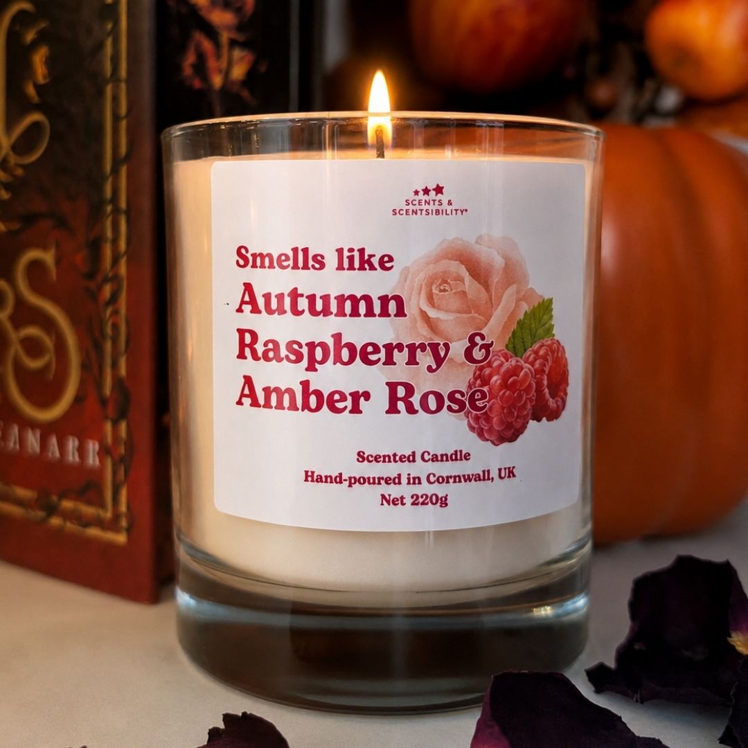 A lit scented candle in a clear glass jar with a white label reading 'Smells like Autumn Raspberry & Amber Rose.' The candle is photographed on a tabletop with pumpkins and dried petals in the background, creating a seasonal setting.