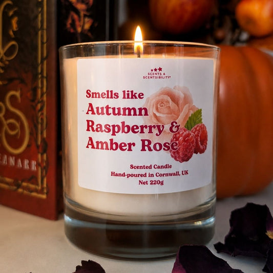 A lit scented candle in a clear glass jar with a white label reading 'Smells like Autumn Raspberry & Amber Rose.' The candle is photographed on a tabletop with pumpkins and dried petals in the background, creating a seasonal setting.