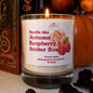 A lit scented candle in a clear glass jar with a white label reading 'Smells like Autumn Raspberry & Amber Rose.' The candle is photographed on a tabletop with pumpkins and dried petals in the background, creating a seasonal setting.