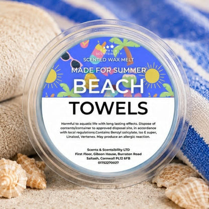 A round clear plastic tub of Scents & Scentsibility Beach Towels scented wax melt, surrounded by seashells and a blue-and-white striped towel on sand.