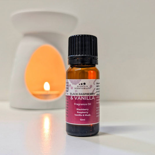 A 10ml bottle of Scents & Scentsibility Black Raspberry & Vanilla Fragrance Oil stands in the foreground on a white surface, with a softly blurred white wax warmer glowing in the background.