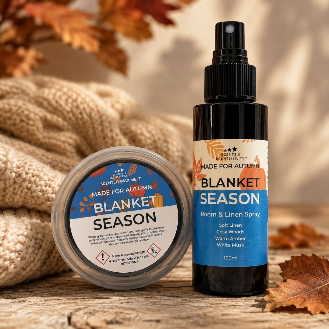 A bottle of Blanket Season room and linen spray next to a round tin of the same scent, styled on a rustic wooden surface with a knitted blanket and autumn leaves in the background.