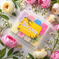 Set of colourful wax melts in a clear container with 'Bloom' branding, surrounded by flowers on a light background.