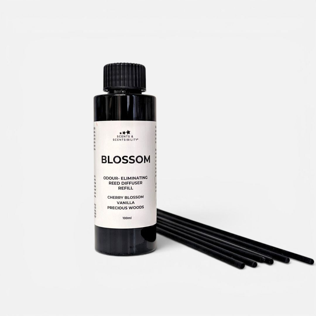 A black bottle of Blossom odour-eliminating Reed Diffuser Refill with a white label, shown next to several black reeds on a light gray background.