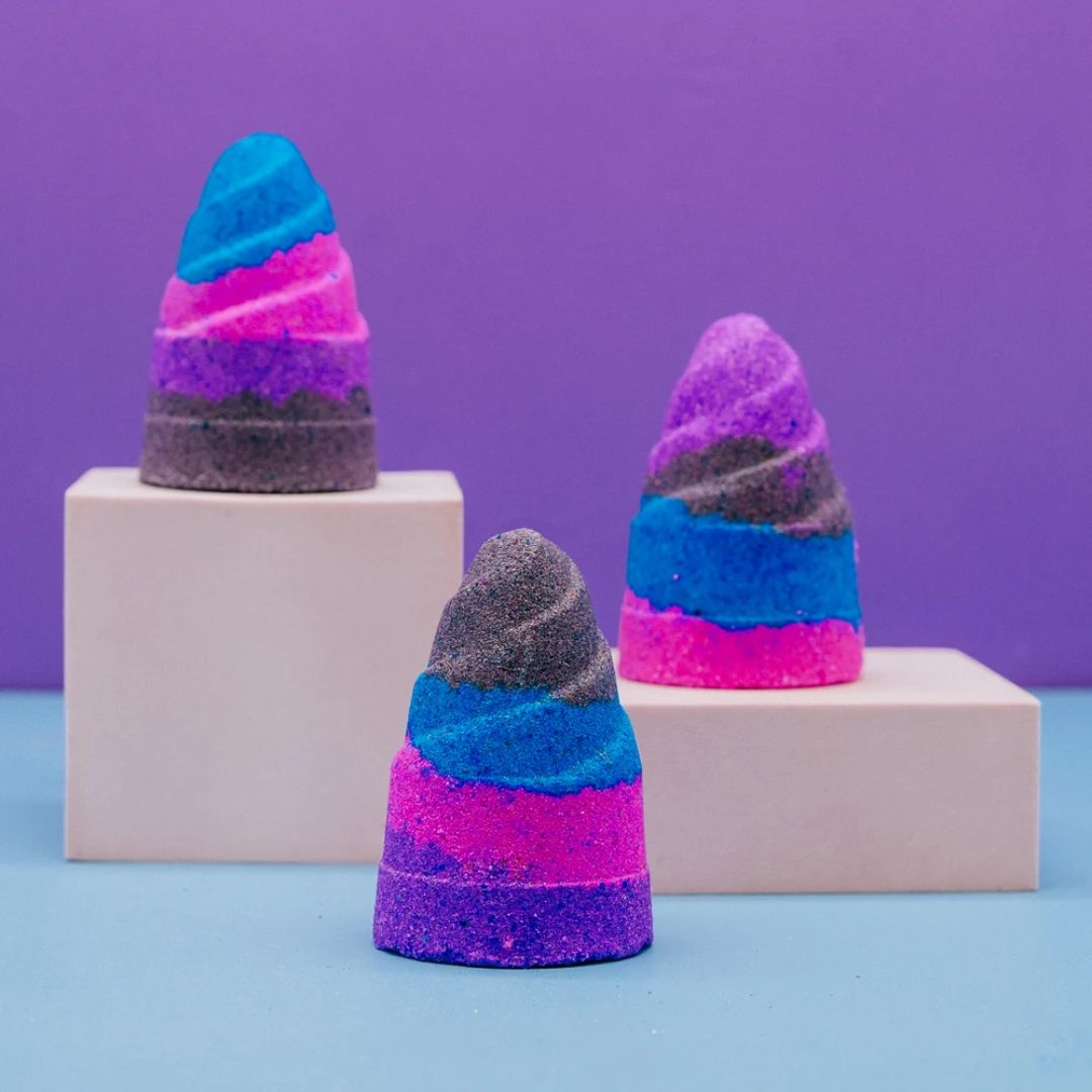 Three cone-shaped bath bombs made of layered glittery powder in blue, pink, purple, and gray, displayed on white pedestals against a purple and light blue background.
