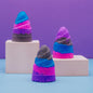 Three cone-shaped bath bombs made of layered glittery powder in blue, pink, purple, and gray, displayed on white pedestals against a purple and light blue background.