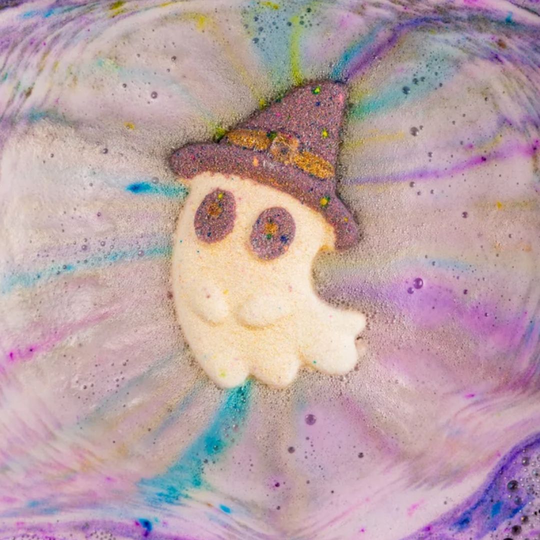 A close-up of a Halloween-themed bath bomb design featuring a cute ghost with a purple witch hat, surrounded by colorful pastel fizzing foam in purple, pink, blue, and white.