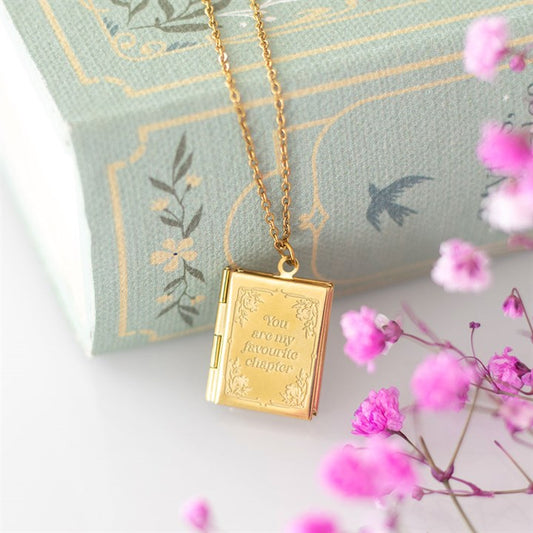Gold book-shaped locket necklace on a decorative pillow with pink flowers.