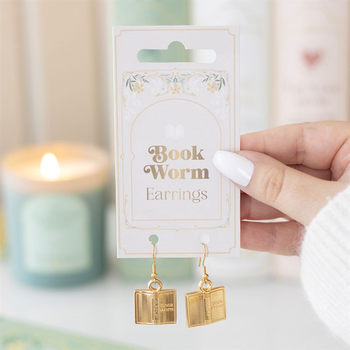 Gold book-shaped earrings on a 'Book Worm' card held by a hand with a blurred candle in the background.