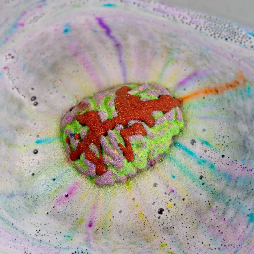 A colorful bath bomb shaped like a brain, featuring a textured lime green and lavender base with orange-red glittery drizzle, resting in a bowl of white foam with pastel rainbow dye splatters.