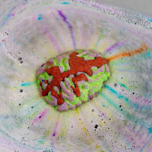 A colorful bath bomb shaped like a brain, featuring a textured lime green and lavender base with orange-red glittery drizzle, resting in a bowl of white foam with pastel rainbow dye splatters.