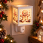 A white square plug-in wax melt warmer with a warm glowing LED panel featuring a gingerbread man illustration, shown mounted on a wall next to Christmas decorations.