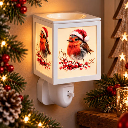 A white cube-shaped Christmas plug-in wax warmer with a warm glowing face and printed robin bird illustrations wearing Santa hats, shown lit in a holiday setting with pine branches, red berries, and warm lights.