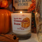 A lit scented candle in a clear glass jar labeled 'Smells like Cinnamon Buns' by Scents & Scentsibility, styled with pumpkins, cinnamon sticks, and a decorative vase in a warm autumn setting.