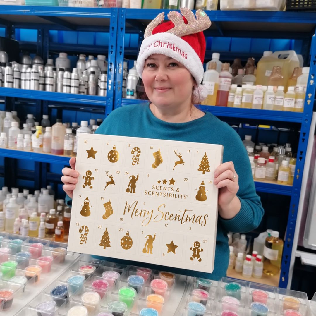 Clare wearing a Santa hat, holding a giant white wax melt advent calendar, in front of shelves filled with fragrance oils and a table full of wax melts. 