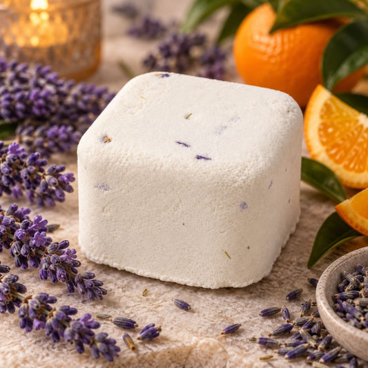 A square white shower steamer with visible lavender buds embedded in the surface, styled on a textured beige surface with lavender sprigs, orange slices, green leaves, and a small bowl of lavender.
