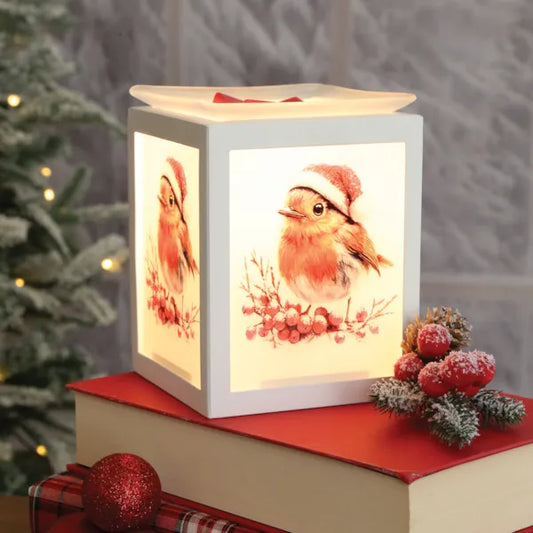 A square white Christmas wax melt warmer with a warm glow, featuring painted robin birds wearing Santa hats on the front panels. The lamp is styled on top of a red book with festive holiday decorations, including a red ornament and frosted berries, with a blurred Christmas tree in the background.