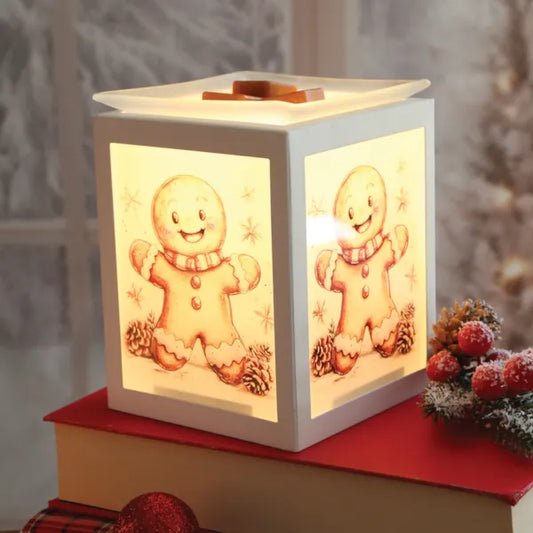 A square white Christmas wax melt warmer with glowing warm panels showing gingerbread men and snowflake designs, styled on a red book with holiday decorations in a cozy indoor setting.