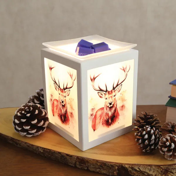 A cube-shaped white wax warmer lamp with watercolor deer illustrations on the front panels, glowing with warm light. A small wax melt sits on the top lid, and the lamp is styled on a wooden slice with pinecones against a neutral background.