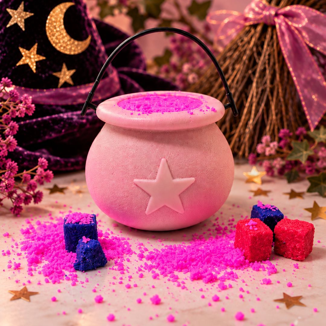 A pink cauldron-shaped bath bomb in a decorative pot with a star emblem on the front, filled with bright pink powder and surrounded by matching pink crystals. The scene is styled with witch-themed props including a purple hat with gold stars, a broom, and small red and blue cubes on a tabletop.