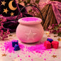 A pink cauldron-shaped bath bomb in a decorative pot with a star emblem on the front, filled with bright pink powder and surrounded by matching pink crystals. The scene is styled with witch-themed props including a purple hat with gold stars, a broom, and small red and blue cubes on a tabletop.