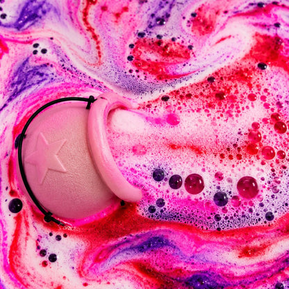 A pink cauldron shaped bath bomb with a star detail, surrounded by vibrant pink, red, and purple foaming bath water with bubbles.