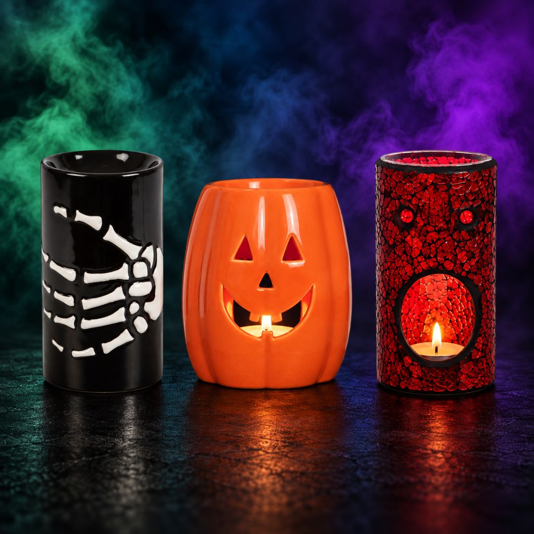 Three Halloween-themed wax melt burners displayed on a dark reflective surface with smoky colored background lighting. Left holder is black with a white skeleton hand design; center holder is orange with a jack-o'-lantern face and lit candle inside; right holder is red with a cracked pattern and a circular cutout showing a lit candle.