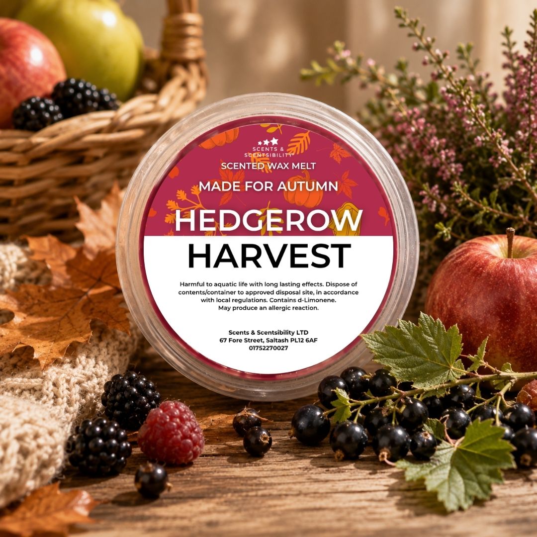 A round plastic tub of scented wax melt labeled 'HEDGEROW HARVEST' sits on a rustic wooden surface surrounded by autumn leaves, apples, blackberries, raspberries, and flowers. The label is visible and the product is clearly the wax melt container.
