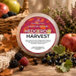 A round plastic tub of scented wax melt labeled 'HEDGEROW HARVEST' sits on a rustic wooden surface surrounded by autumn leaves, apples, blackberries, raspberries, and flowers. The label is visible and the product is clearly the wax melt container.