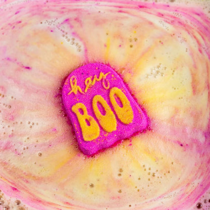 A close-up of a bright pink bath bomb with yellow lettering that reads 'hey BOO', placed in swirling pink and pale yellow foamy water.