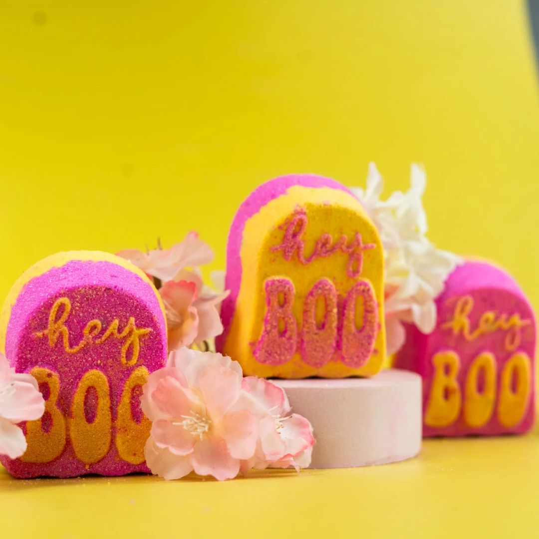 Three tombstone-shaped bath bombs with 'hey BOO' written on them, styled with pink flowers on a bright yellow background.