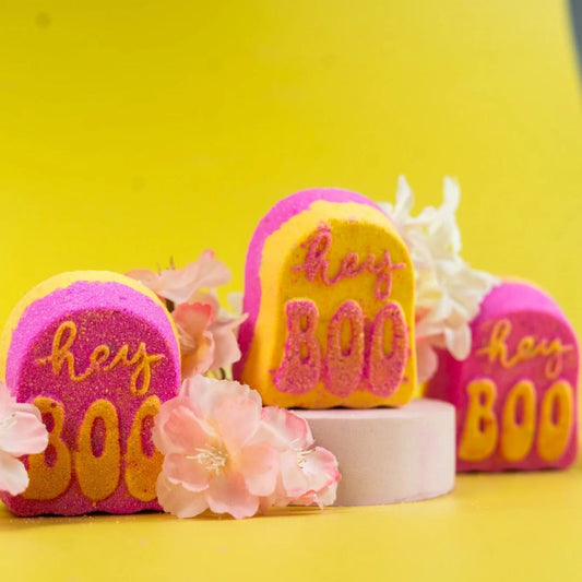 Three tombstone-shaped bath bombs with 'hey BOO' written on them, styled with pink flowers on a bright yellow background.