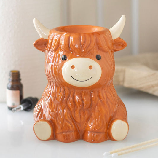 Highland Cow Ceramic Wax Melt and Oil Burner displayed on a white surface with fragrance oil and matches nearby, showing its cute ginger Highland cow design and built-in warming dish.