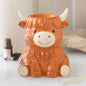 Highland Cow Ceramic Wax Melt and Oil Burner displayed on a white surface with fragrance oil and matches nearby, showing its cute ginger Highland cow design and built-in warming dish.