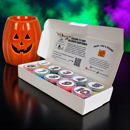 An open white gift box containing multiple small pots of colorful wax melts, with a printed insert card inside the lid. A carved orange jack-o'-lantern candle holder sits beside the box on a dark reflective surface, with a blurred green and purple festive background.