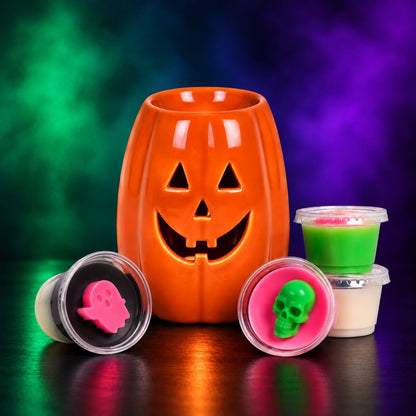 A Halloween-themed wax melt set featuring a glowing orange jack-o'-lantern-shaped wax melt burner in the center, with three small clear plastic pots containing pink, green, and white wax melts arranged around it on a reflective dark surface against a smoky multicolor background.