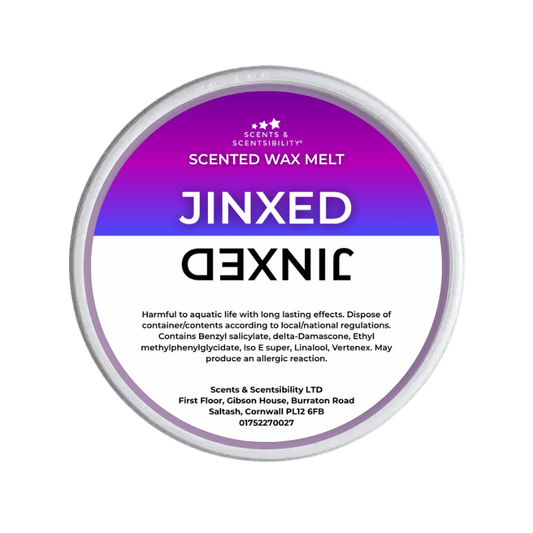 A Jinxed Scented Wax Melt by Scents & Scentsibility, featuring a purple-to-white gradient label with bold white text, shown against a solid dark green background.