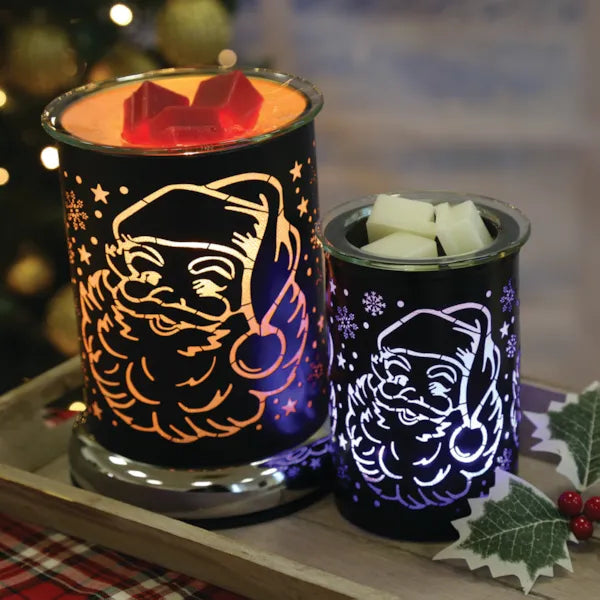 Two decorative wax warmers with Santa Claus designs on a table with a blurred Christmas tree background.