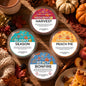 Four round scented wax melts in clear plastic tubs arranged on a rustic wooden surface with autumn-themed props including pumpkins, leaves, cinnamon, and a bowl of tea. The labels read Hedgerow Harvest, Blanket Season, Pumpkin Peach Pie, and Beach Bonfire.