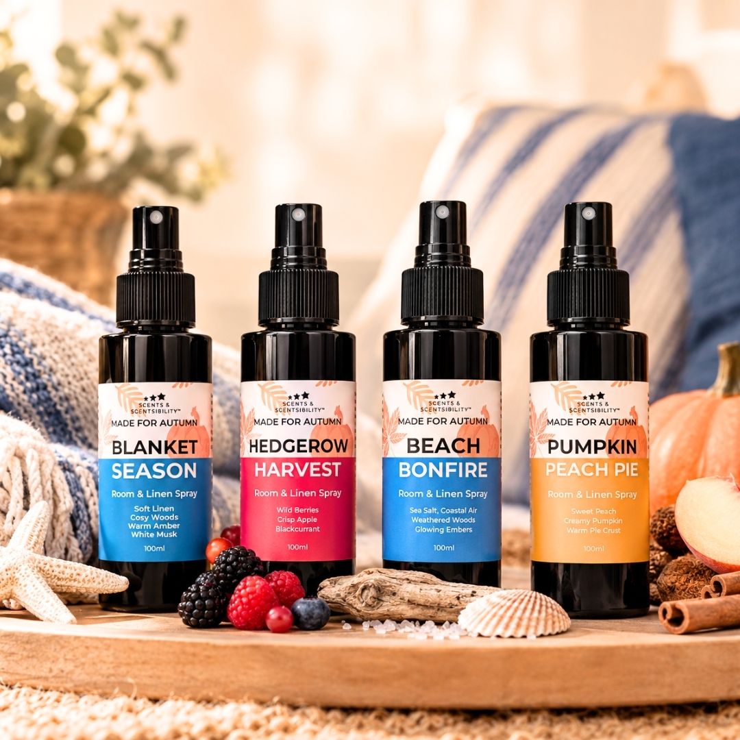 Four fragrance spray bottles labeled Blanket Season, Hedgewood Harvest, Beach Bonfire, and Pumpkin Peach Pie, arranged on a wooden surface with autumn-themed props including berries, starfish, shells, and a blue striped blanket in the background.