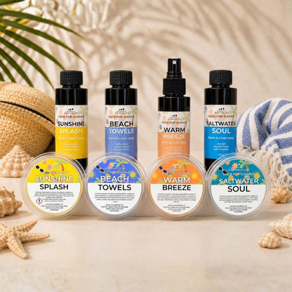 A set of four scented products arranged on a light beige surface with beach-themed props: two spray bottles labeled Sunshine and Warm Breeze, two pump bottles labeled Beach Towels and Saltwater Soul, and four round wax melt tubs labeled Sunshine Splash, Beach Towels, Warm Breeze, and Saltwater Soul. The scene includes a straw hat, starfish, shells, and a blue-and-white towel.