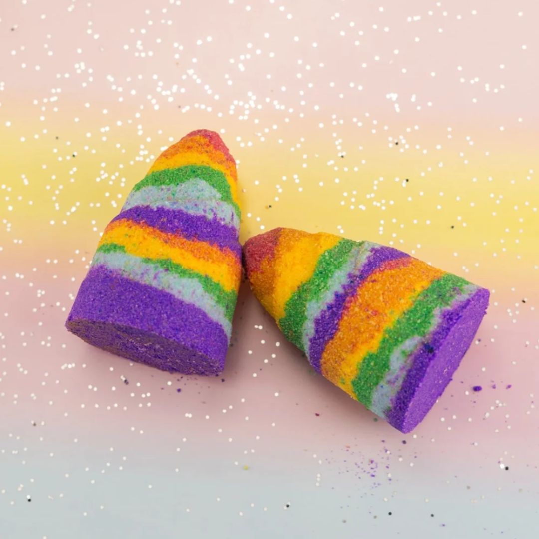 Two cone-shaped bath bombs with rainbow-colored stripes of purple, orange, green, yellow, and pink, decorated with glitter, set against a pastel gradient background with white glitter specks.