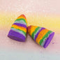 Two cone-shaped bath bombs with rainbow-colored stripes of purple, orange, green, yellow, and pink, decorated with glitter, set against a pastel gradient background with white glitter specks.